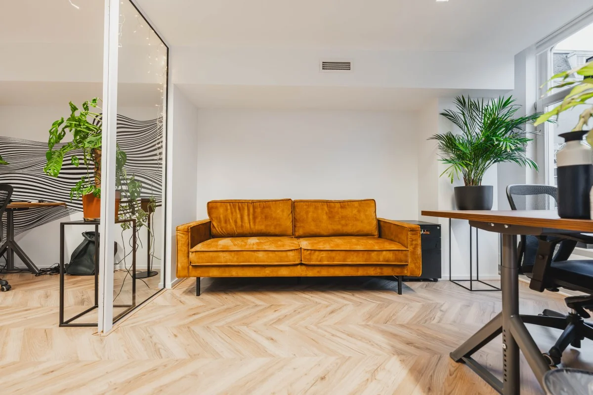 Moderne kantoorruimte aan de Korte Reguliersdwarsstraat met een okergele bank, houten visgraatvloer en groene kamerplanten.